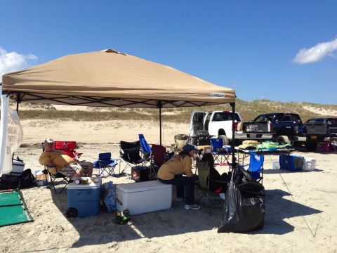 1979 Ford Bronco - People relax under a canopy on a beach with several trucks parked nearby.