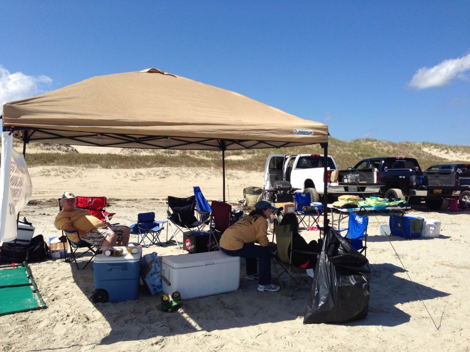 Several pickup trucks parked on a sandy beach under a tan canopy with people sitting in chairs