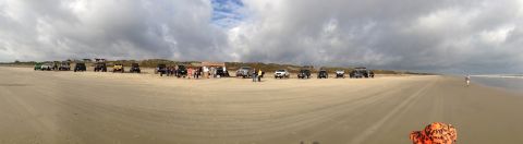 1979 Ford Bronco - Several off-road vehicles are gathered on a wide, sandy beach under a dramatic, cloudy sky.