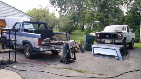 1983 Ford Bronco - Two Ford Broncos, a blue one with its hood open and a white one with its camper shell removed, sit in a driveway next to a welding machine.