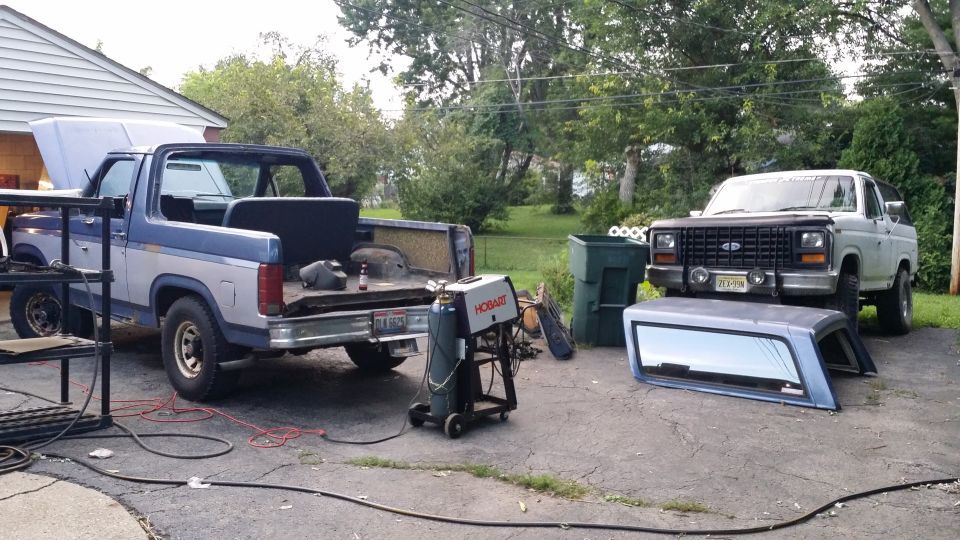 Two 1980s Ford Broncos in a driveway, one blue with hood up, one white with camper shell removed