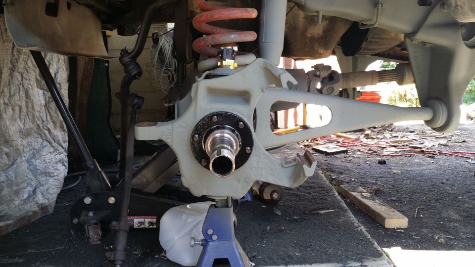 Close-up of vehicle's front suspension assembly, including the spindle, brake rotor area, coil spring, and control arms. Vehicle is on jack stands