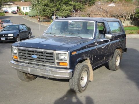 1985 Ford Bronco - A dirt-covered dark blue Ford Bronco is parked on a street, viewed from the front three-quarter angle.