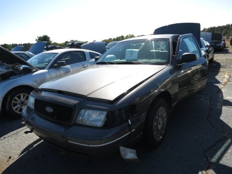 2001 Ford Crown Victoria - A dark gray 2001 Ford Crown Victoria sits in a lot, its front end showing damage and a detached turn signal.