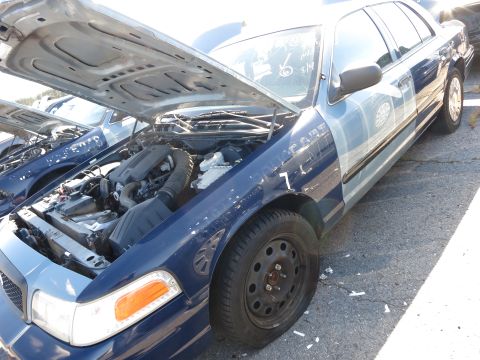 2001 Ford Crown Victoria - A blue Ford Crown Victoria police car is shown with its hood open, revealing the engine compartment. Another similar vehicle is visible to…
