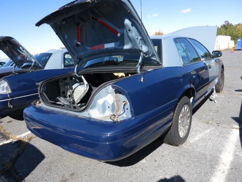 2001 Ford Crown Victoria - Two blue Ford Crown Victorias are parked outside, both with hoods and one with the trunk open.