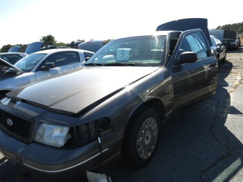 2001 Ford Crown Victoria - A dark sedan sits in a junkyard with its hood and trunk open, alongside other vehicles under a clear sky.