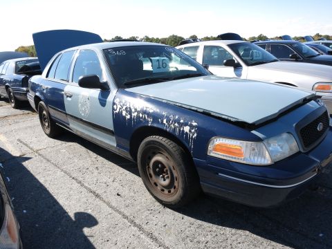 2001 Ford Crown Victoria - A blue and light blue 2001 Ford Crown Victoria sits in a lot with other vehicles, its hood open and paint showing wear.