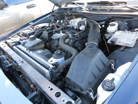2001 Ford Crown Victoria - Close-up of a car's engine bay, showing various mechanical parts and hoses. Another vehicle is partially visible behind.