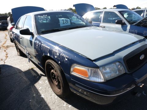 2001 Ford Crown Victoria - A damaged 2001 Ford Crown Victoria sits in a junkyard with its hood open and other vehicles in the background.