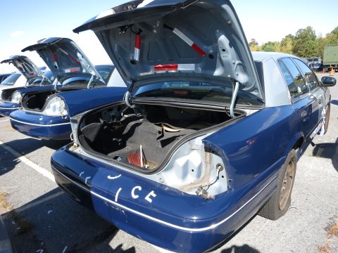 2001 Ford Crown Victoria - Multiple blue and silver police cars are parked in a lot, with their trunks open. The car in the foreground has the partial word 'ICE' visi…