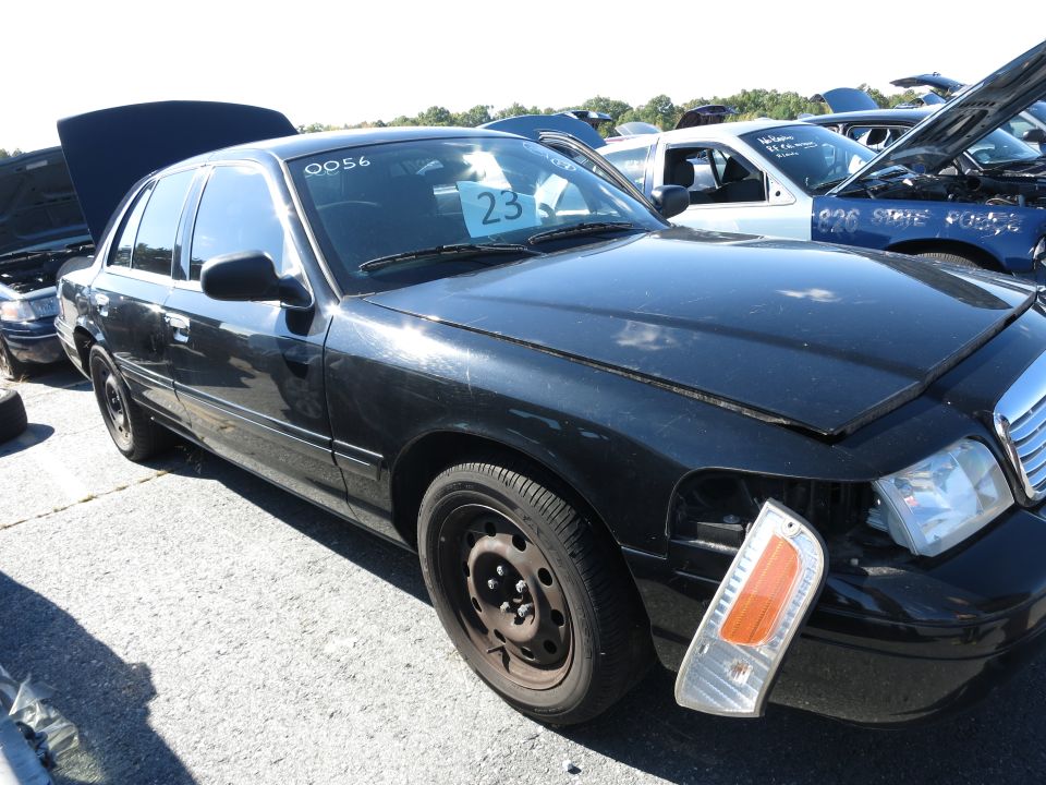 Black sedan with missing headlight assembly and a number 23 sign on the windshield. Other cars with hoods up in background