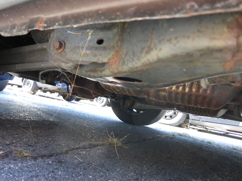 Underside of a rusty vehicle, showing frame and exhaust system. Multiple vehicles visible in background