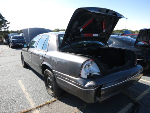 2001 Ford Crown Victoria - A dark gray 2001 Ford Crown Victoria sits in a lot with its trunk and hood open, showing a flat rear tire and damaged taillight.