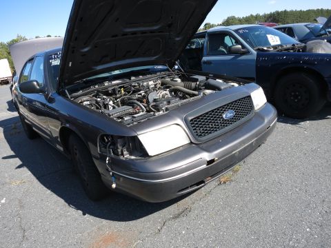 2001 Ford Crown Victoria - A dark gray 2001 Ford Crown Victoria is parked with its hood open, showing the engine compartment and a damaged front bumper.
