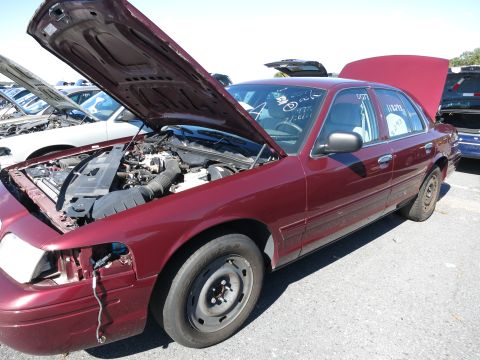 2001 Ford Crown Victoria - A maroon 2001 Ford Crown Victoria sedan sits in a junkyard with its hood open, revealing the engine compartment.