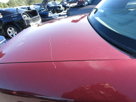 2001 Ford Crown Victoria - A close-up of a red car's hood and windshield shows scratches, with other vehicles visible in the background of a junkyard.