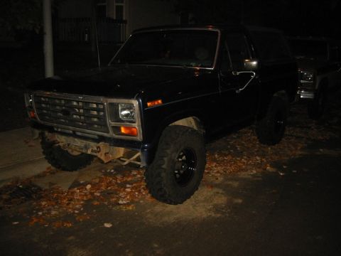 1985 Ford Bronco - A dark Ford Bronco is parked outside at night with fallen leaves on the ground. Another vehicle is visible behind it.