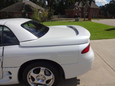 1995 Mitsubishi 3000GT - The rear of a white 1995 Mitsubishi 3000GT is visible, parked on a concrete driveway with houses in the background.