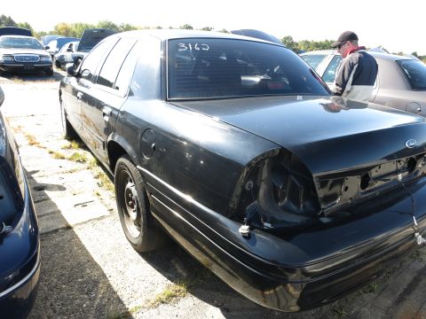 2001 Ford Crown Victoria - Rear view of a black 2001 Ford Crown Victoria sedan in a junkyard, showing damage to the rear bumper and taillight assembly.
