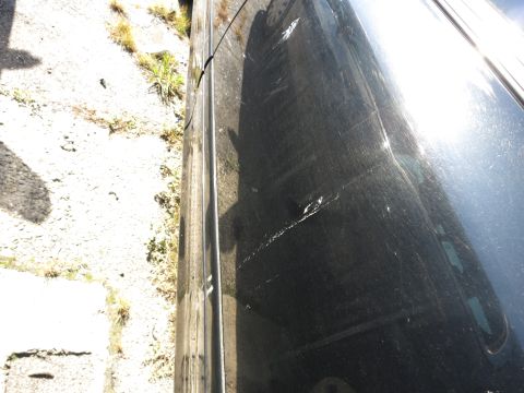 2001 Ford Crown Victoria - A black car door shows significant damage, including scratches and a dent, under bright sunlight.
