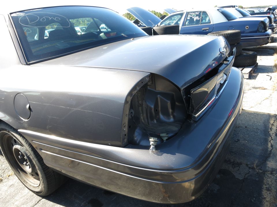 Rear quarter view of a gray 2001 Ford Crown Victoria in a junkyard. Other cars are visible in the background