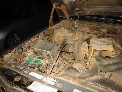 1985 Ford Bronco - The engine bay of a vehicle, showing the battery and various hoses and components. Another vehicle is partially visible in the background.