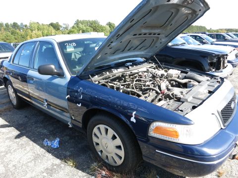 2001 Ford Crown Victoria - A two-tone blue and white Ford Crown Victoria has its hood open, revealing the engine compartment in a vehicle yard.