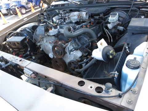 2001 Ford Crown Victoria - The engine bay of a vehicle, showing the V8 engine, various hoses, and fluid reservoirs, with other vehicles visible in the background.