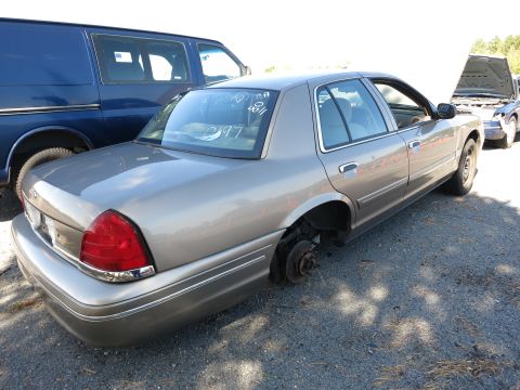 2001 Ford Crown Victoria - A gray 2001 Ford Crown Victoria is seen with its rear wheel removed, parked in a lot with other vehicles.