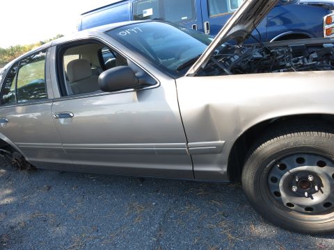 2001 Ford Crown Victoria - A damaged 2001 Ford Crown Victoria sits in a junkyard with its hood open, next to a blue van.