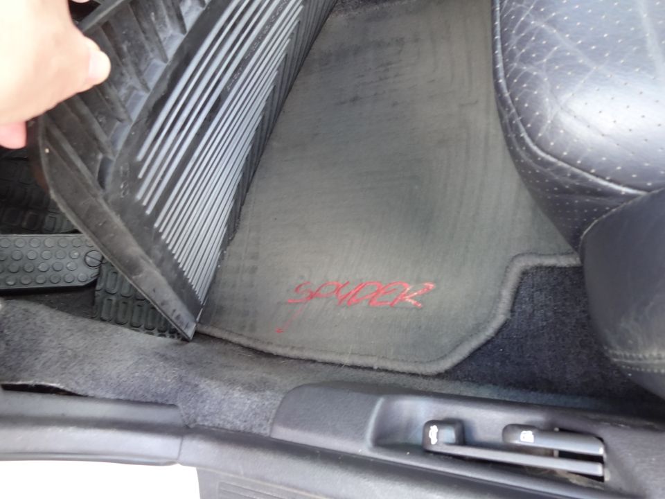 Car floor mat with red 'Spyder' lettering, partially obscured by a hand lifting a rubber mat