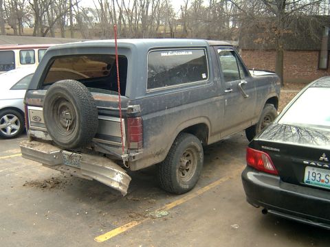 1982 Ford Bronco - This dark blue SUV, believed to be a 1982 Ford Bronco, is parked with its spare tire mounted on the rear and a bent bumper.