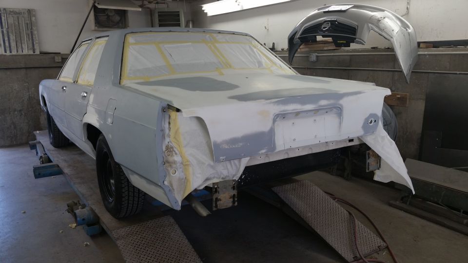 Car bodywork in progress, rear view. The car is on a lift with primer and tape on the body panels