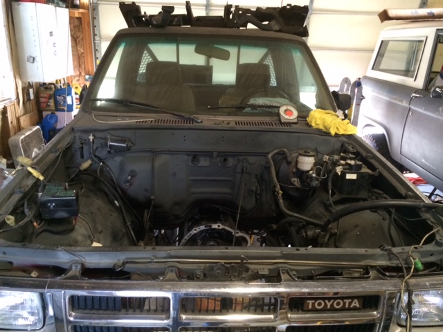 Truck engine bay with transmission removed, showing engine block and brake master cylinder. Another vehicle is parked behind