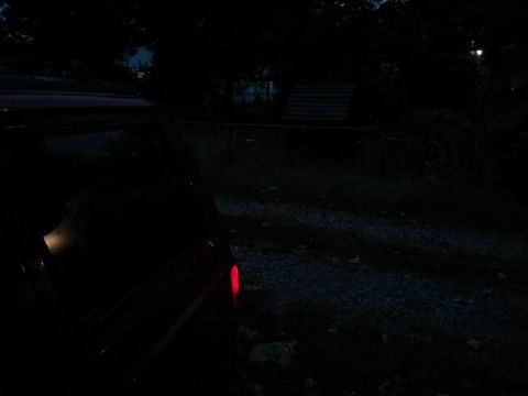 1986 Ford Bronco - A vehicle's rear taillight glows red in the dark, with trees and a fence in the background.