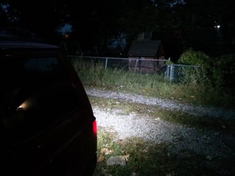 1986 Ford Bronco - The rear of a dark SUV is visible at night, parked on a gravel surface with a fence and shed in the background.