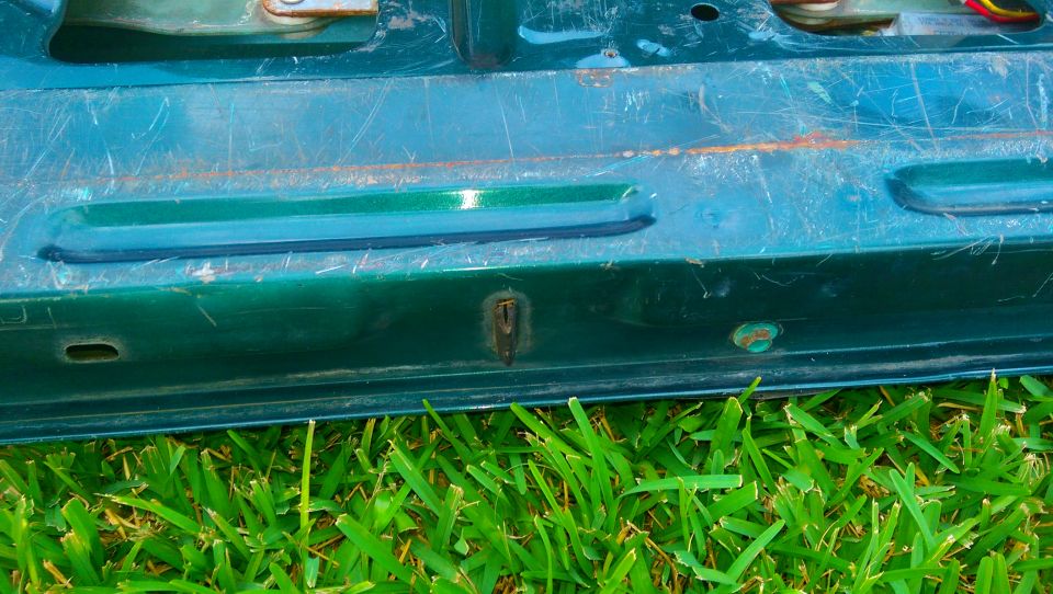 Green tailgate section with visible scratches and a rusty lock mechanism, grass in foreground