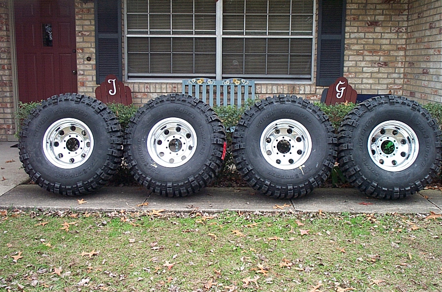 Four mud-terrain tires on polished alloy wheels lined up by brick house