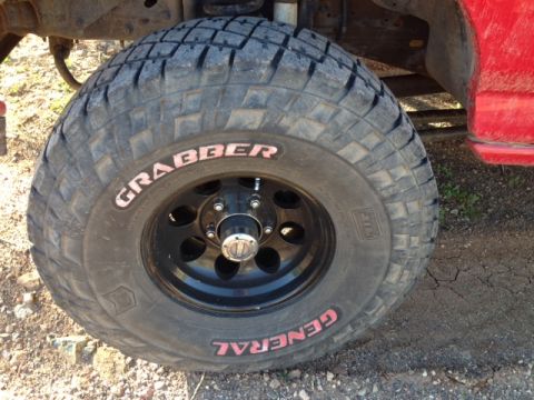 1996 Ford Bronco - A close-up shot focuses on a General Grabber tire mounted on a black wheel, with a portion of a red vehicle in the background.