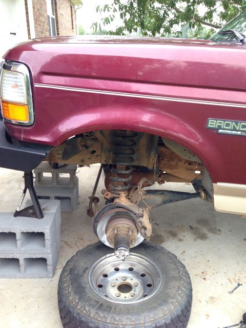 1996 Ford Bronco - The front suspension of a maroon Ford Bronco is visible as it is being worked on, with the wheel removed.
