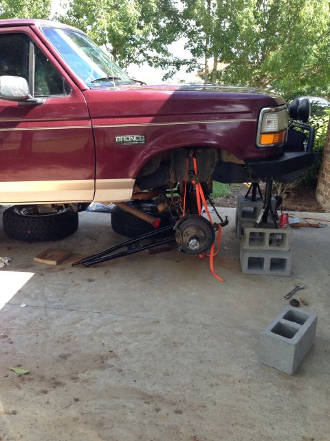 1996 Ford Bronco - Front suspension of a red 1996 Ford Bronco is disassembled and raised on jack stands, indicating a lift kit installation.