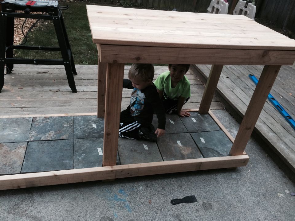 Two young boys are playing under a newly built wooden table on a deck. One boy wears a black shirt with a graphic, the other a green shirt