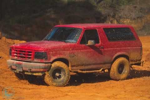 1995 Ford Bronco - A red Ford Bronco navigates a muddy terrain, its body caked in dirt.