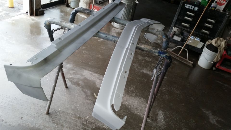 Two car bumper parts, one gray and one white, resting on stands in a workshop