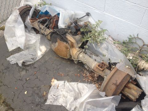 1971 Ford F-250 - A rusty rear axle assembly, likely from a truck, is partially covered by plastic and overgrown with weeds.