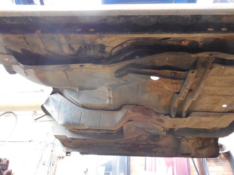 1993 Ford Bronco - Underside of a body tub raised off its frame, showing the rusty stamped floor and toe-board, seat-crossmember reinforcement, open body-moun…