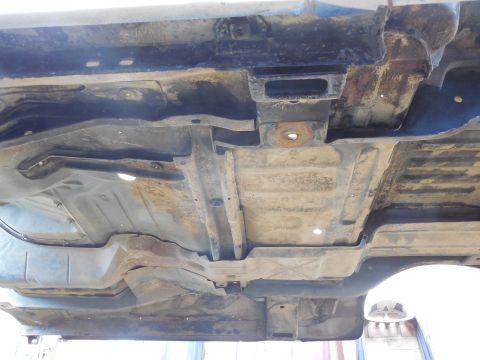 1993 Ford Bronco - Close underside view of a raised body showing the stamped cab floor and toe-board pressings, a body-mount hole with a rusty washer, seat-mo…