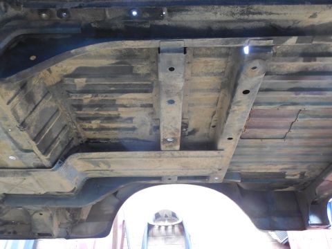 1993 Ford Bronco - Wide underside shot of a lifted body tub showing the stamped ribbed floor pan, wheelhouse, body-mount reinforcement brackets with bolt hole…