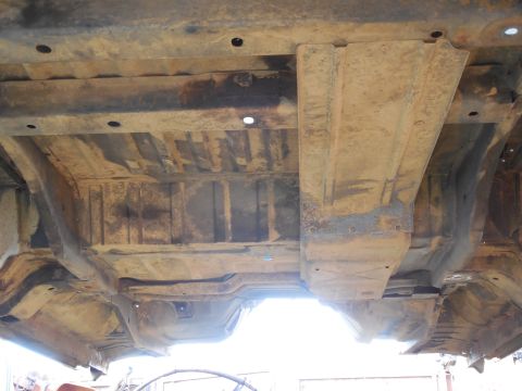 1993 Ford Bronco - Underbody shot of a lifted truck body showing the rusty ribbed floor pan, transmission tunnel hump and a large flat sheet-metal panel spann…